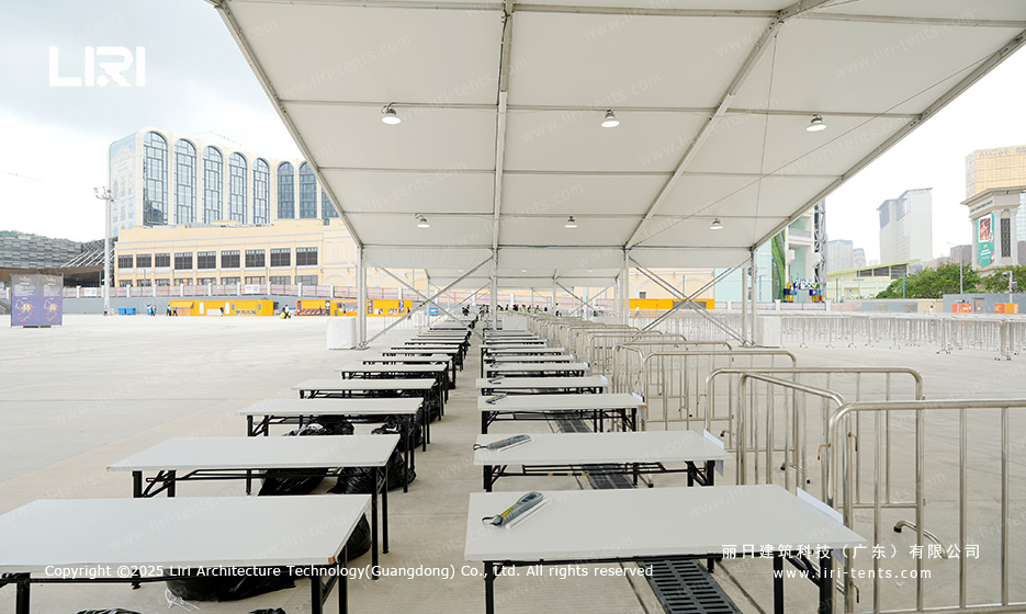 Side aisle of an award ceremony party tent showing long banquet tables and outdoor crowd control barriers in a plaza setting