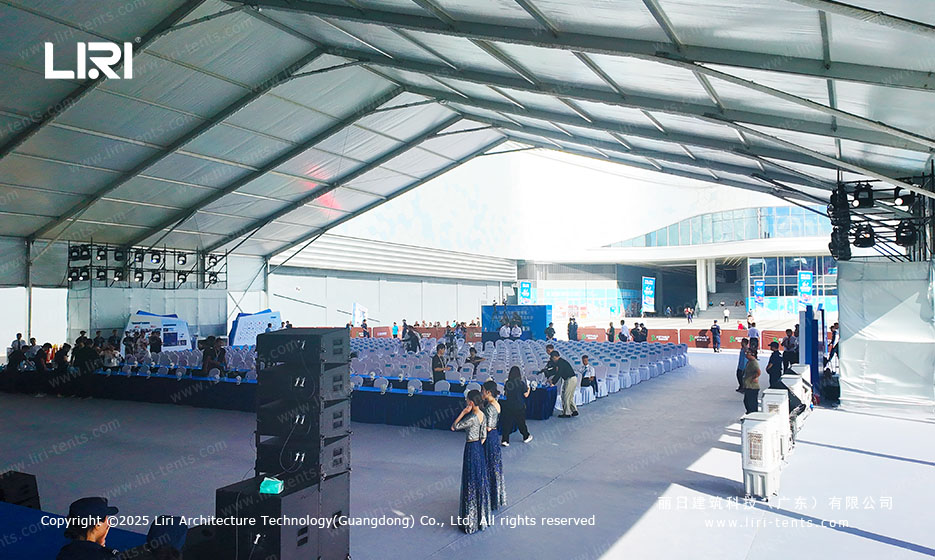 Interior of an opening ceremony party tent showing rows of white-covered chairs and a long blue aisle carpet leading to the stage