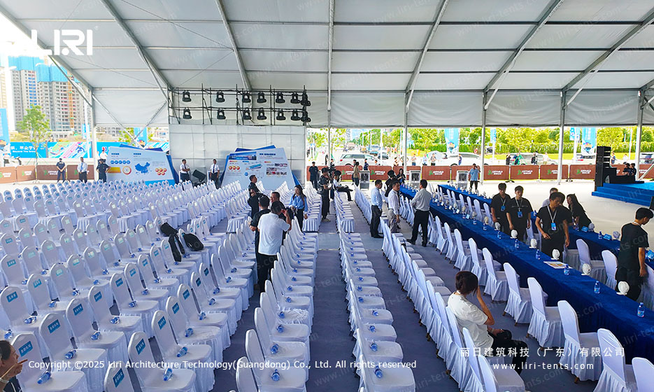 Long blue VIP table running along the side of an opening ceremony party tent with white chair covers and a central aisle carpet