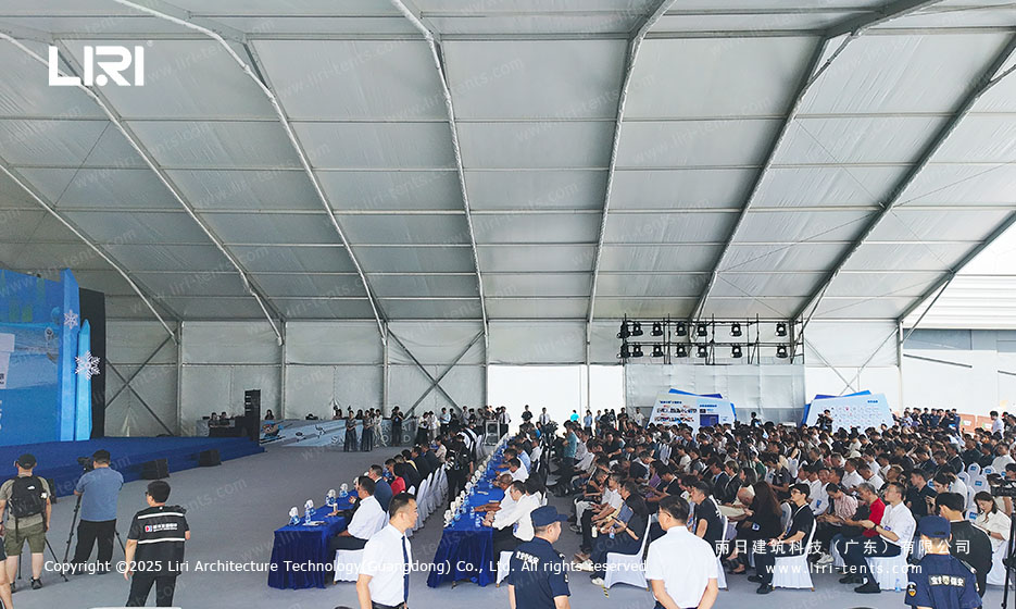 Back half of an opening ceremony party tent with stage, blue backdrop, lighting truss, and a seated audience facing the stage