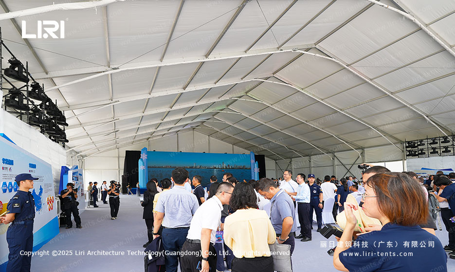 Interior of an opening ceremony party tent with the audience watching the stage, blue aisle carpet, and ceiling truss with stage lighting